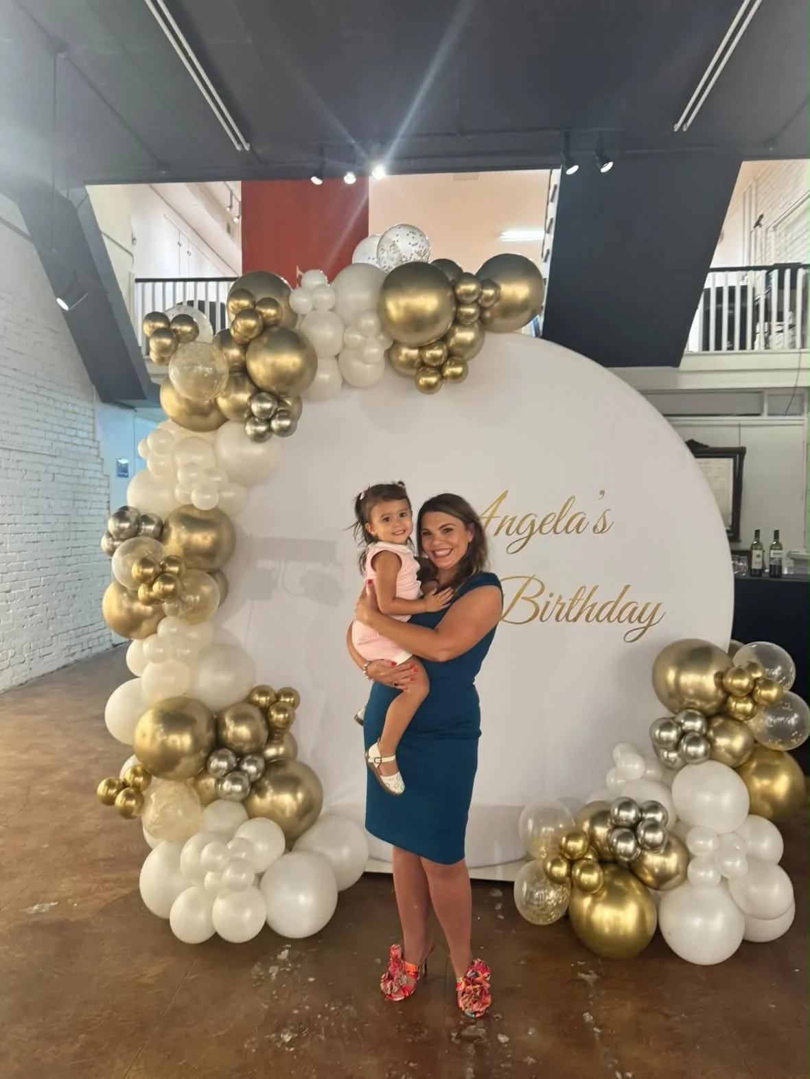 A woman holding a child beside the gold and white birthday balloons.